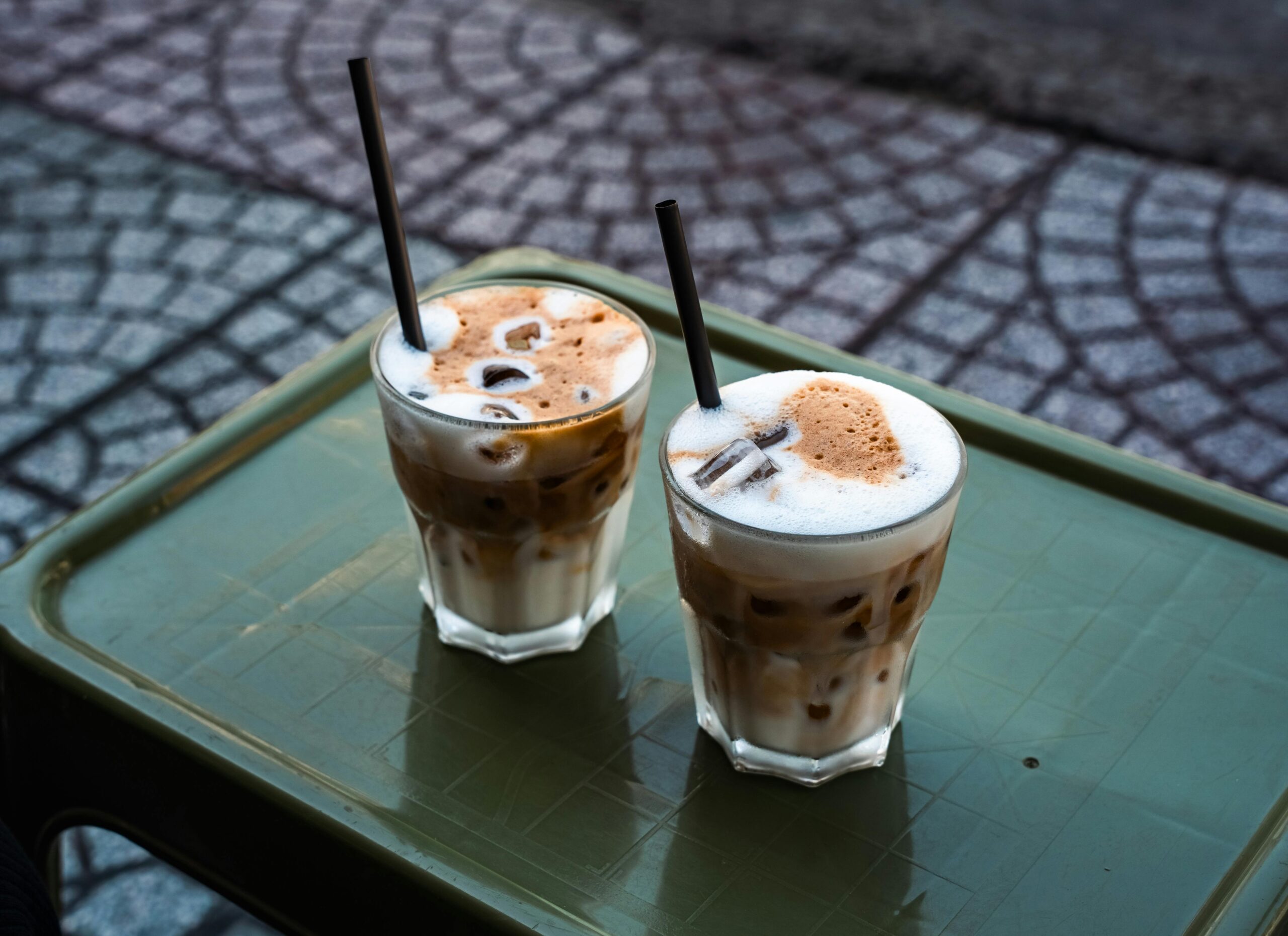 Two iced coffee drinks with straws on a green table outdoors, featuring creamy foam and a refreshing vibe.